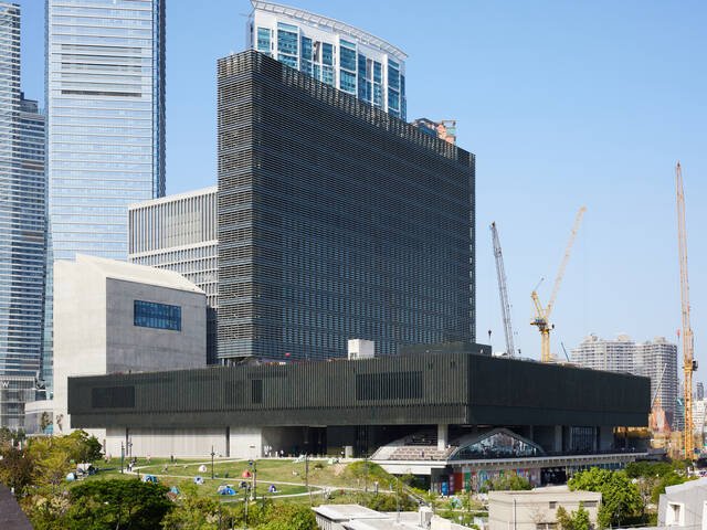 A view of the M+ building, which consists of a square-shaped horizontal podium and a tower stretching across the podium’s centre. From the front, the building looks similar to an upside-down T. It is covered by dark green ceramic panels.